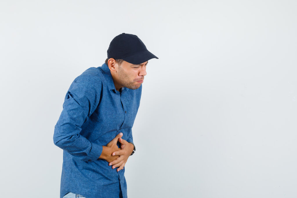 young man in blue shirt, cap suffering from stomach pain and looking unwell , front view.
